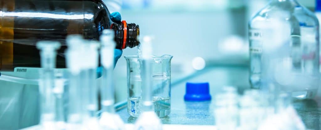Mano con guante vertiendo líquido desde una botella ámbar a un vaso de precipitados en un laboratorio.